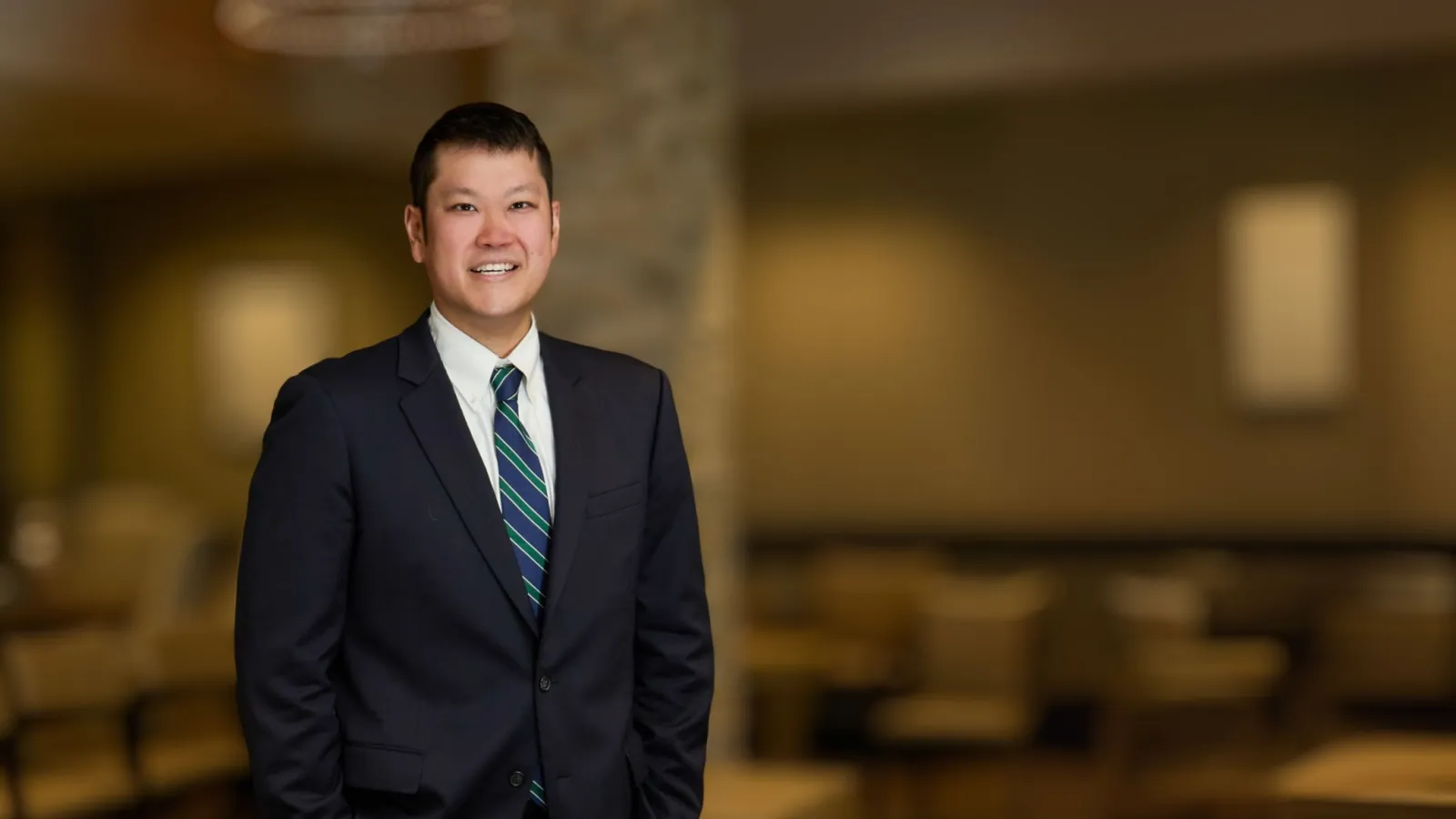 Smiling man in dark suit and striped tie standing in a softly lit modern office or lounge area.