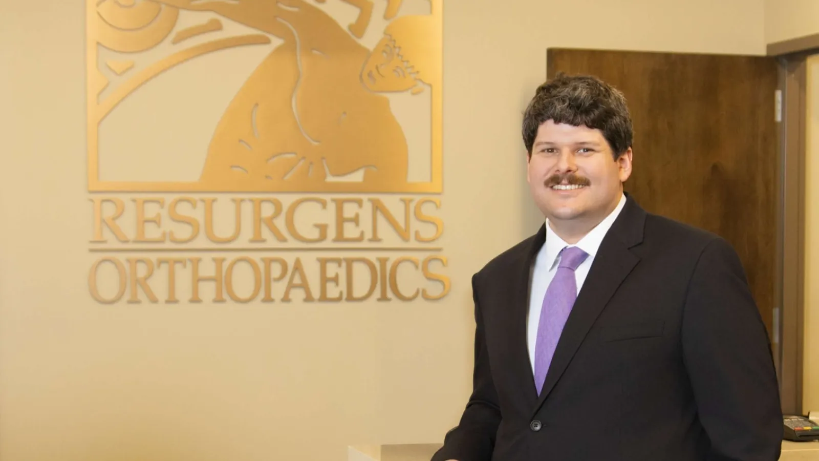 Man in suit stands smiling beside a reception desk with Resurgens Orthopaedics logo on the wall.