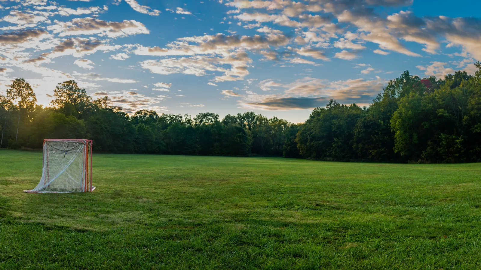 Wide grassy field with a lacrosse goalpost at sunset under a partly cloudy sky and surrounding trees.