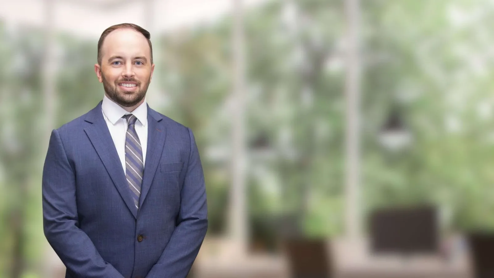 Smiling man in a blue suit and striped tie standing in a bright modern office with large windows and greenery outside