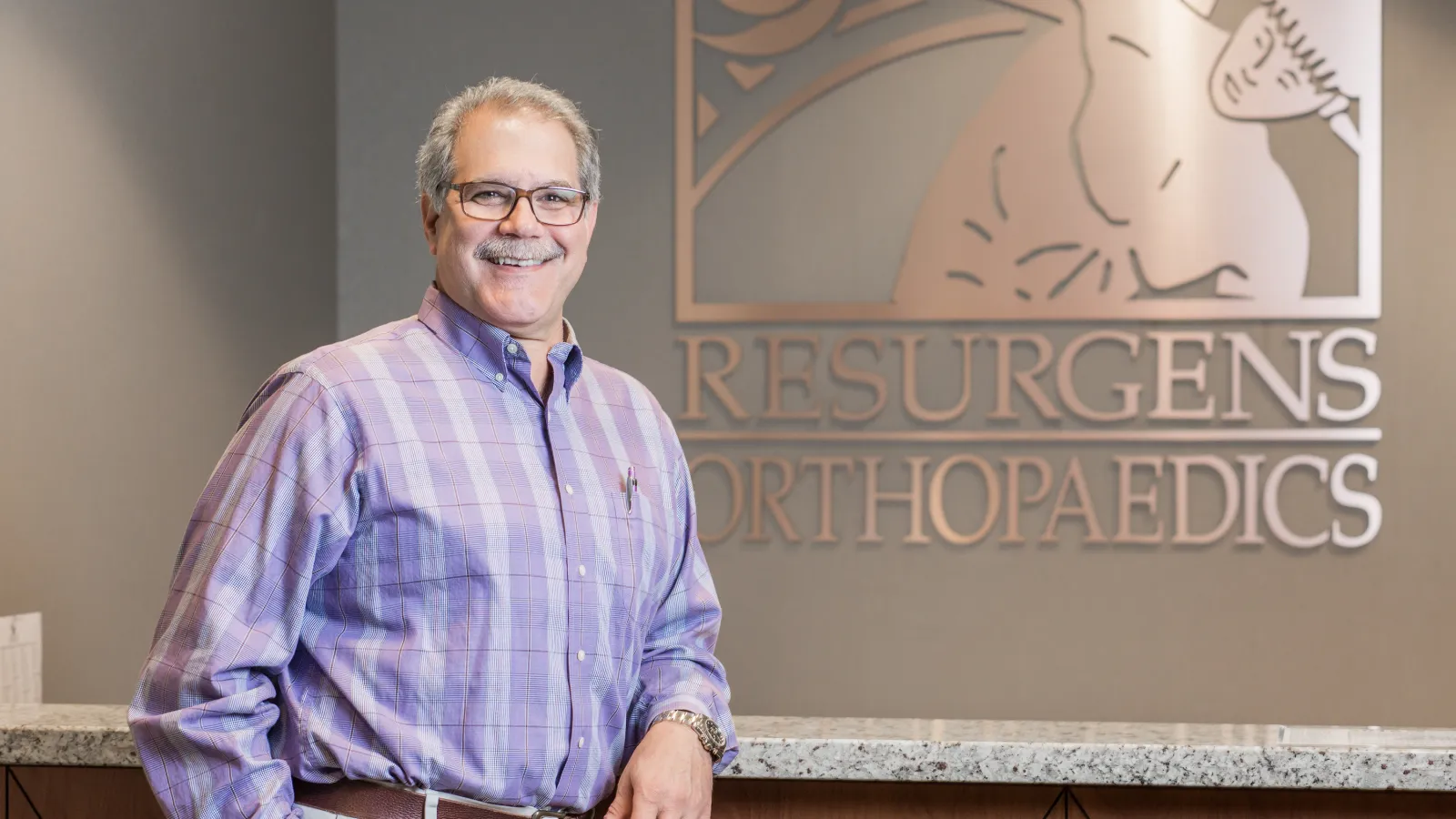 Smiling man in glasses and purple plaid shirt standing at Resurgens Orthopaedics reception desk.
