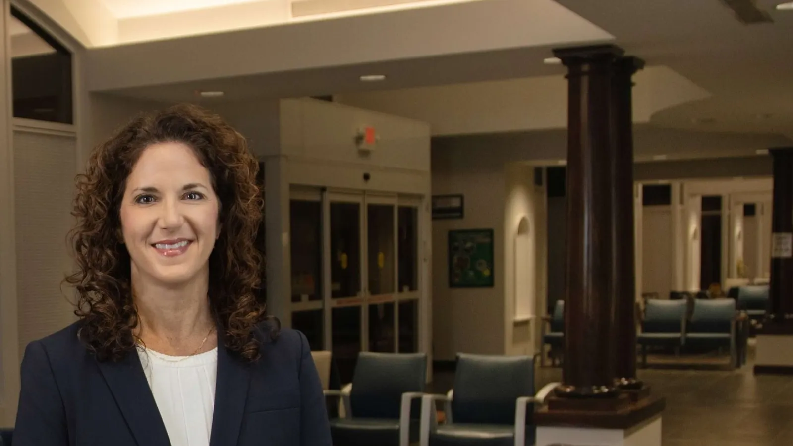 Professional woman with curly hair in navy blazer standing in modern lobby with wooden columns and seating.