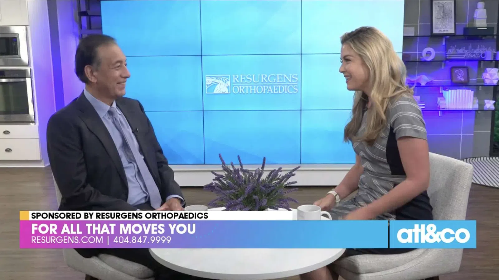 Two people engaged in a studio interview with Resurgens Orthopaedics branding on screen and a flower centerpiece on the table