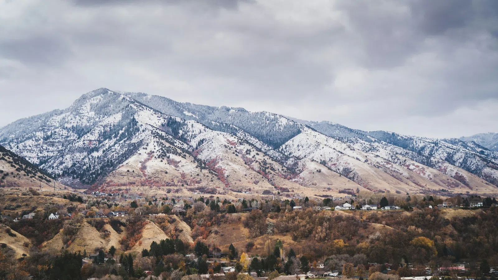 Mountain range with light snow under cloudy sky overlooking autumn-colored forest and scattered houses.
