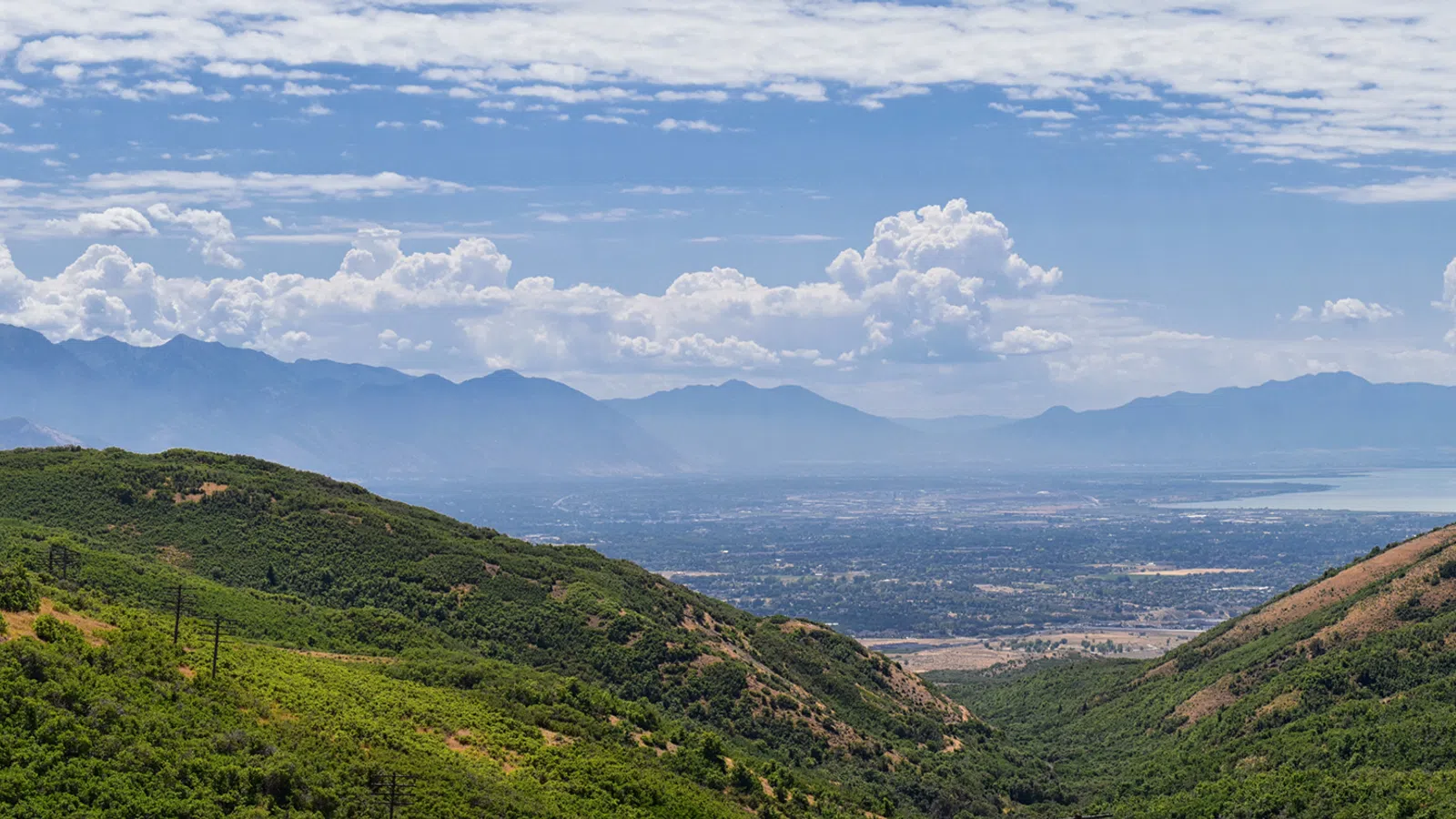 Panoramic view of green hills leading to a distant cityscape with mountains and a partly cloudy sky.