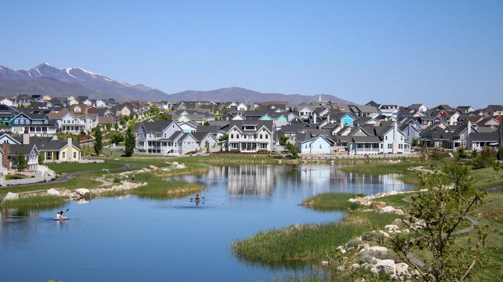 Suburban neighborhood with houses around a calm lake, people kayaking, and mountains in the background on a clear day