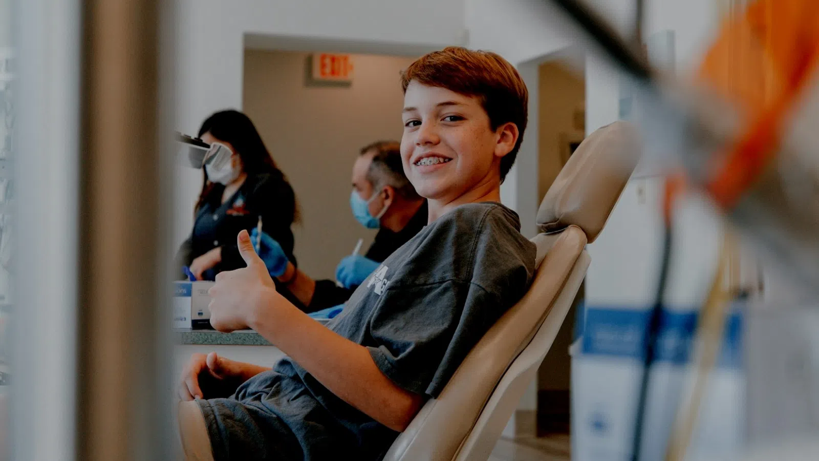 Young boy with braces smiling and giving thumbs up at dental clinic with staff in masks behind him.