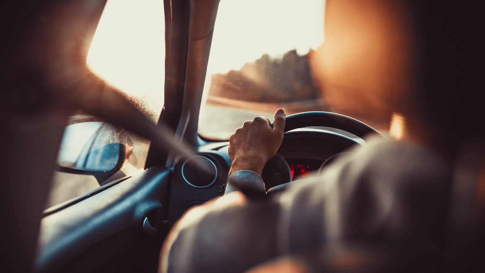 Close-up of a hand gripping a steering wheel, driving into the sunset on a scenic road.