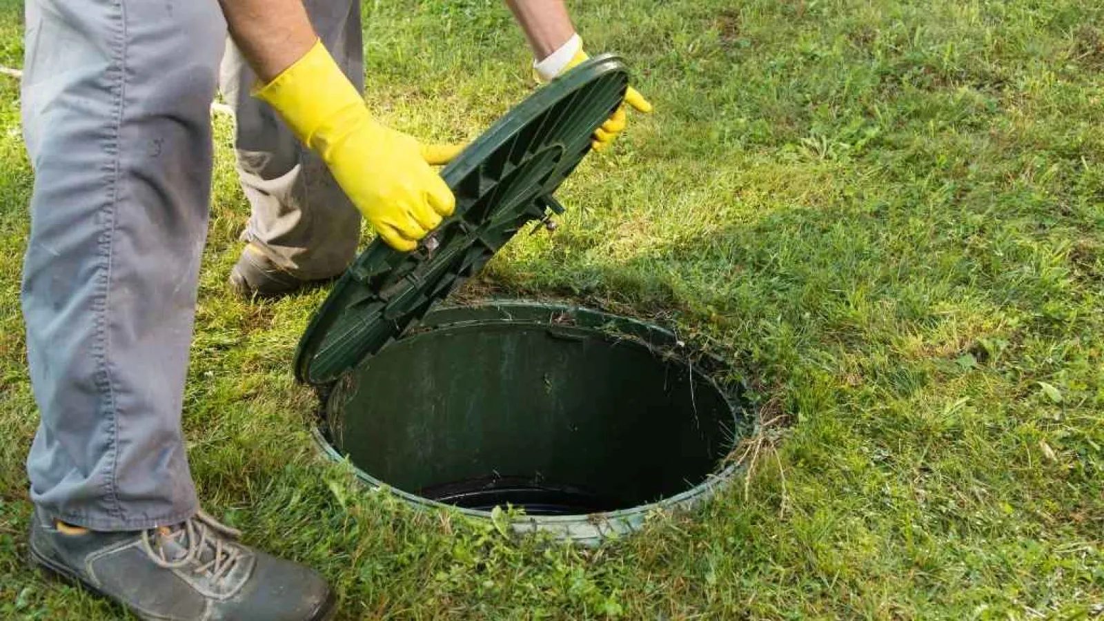 Person wearing yellow gloves lifting a green septic tank lid on grass outdoors.