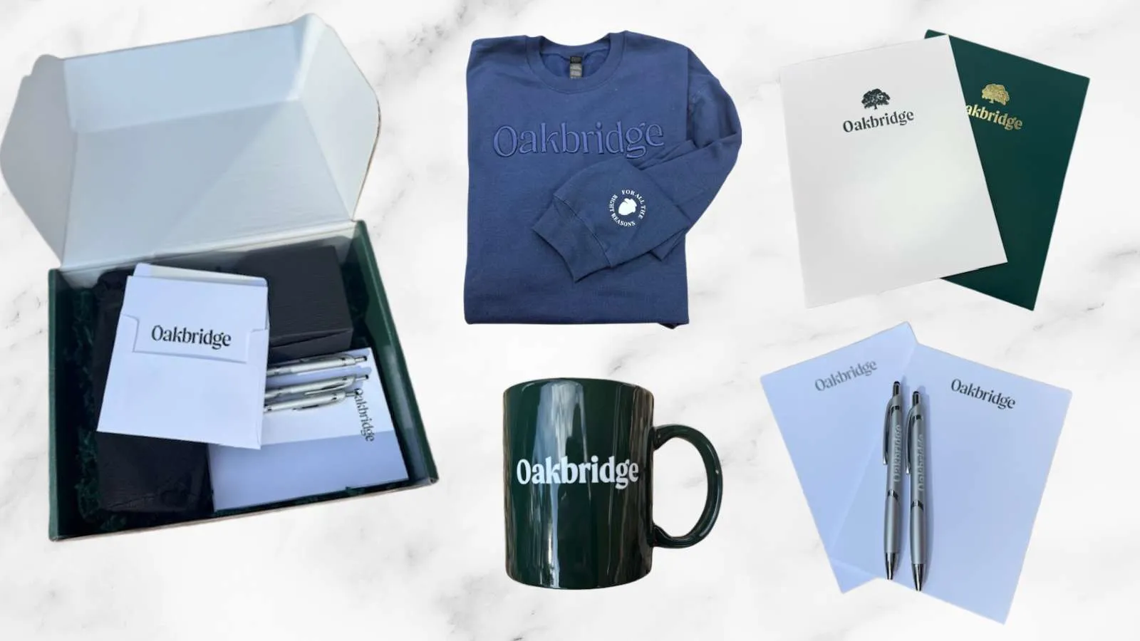 Oakbridge branded merchandise including a navy sweatshirt, black mug, pens, notebooks, and folders on a marble background.