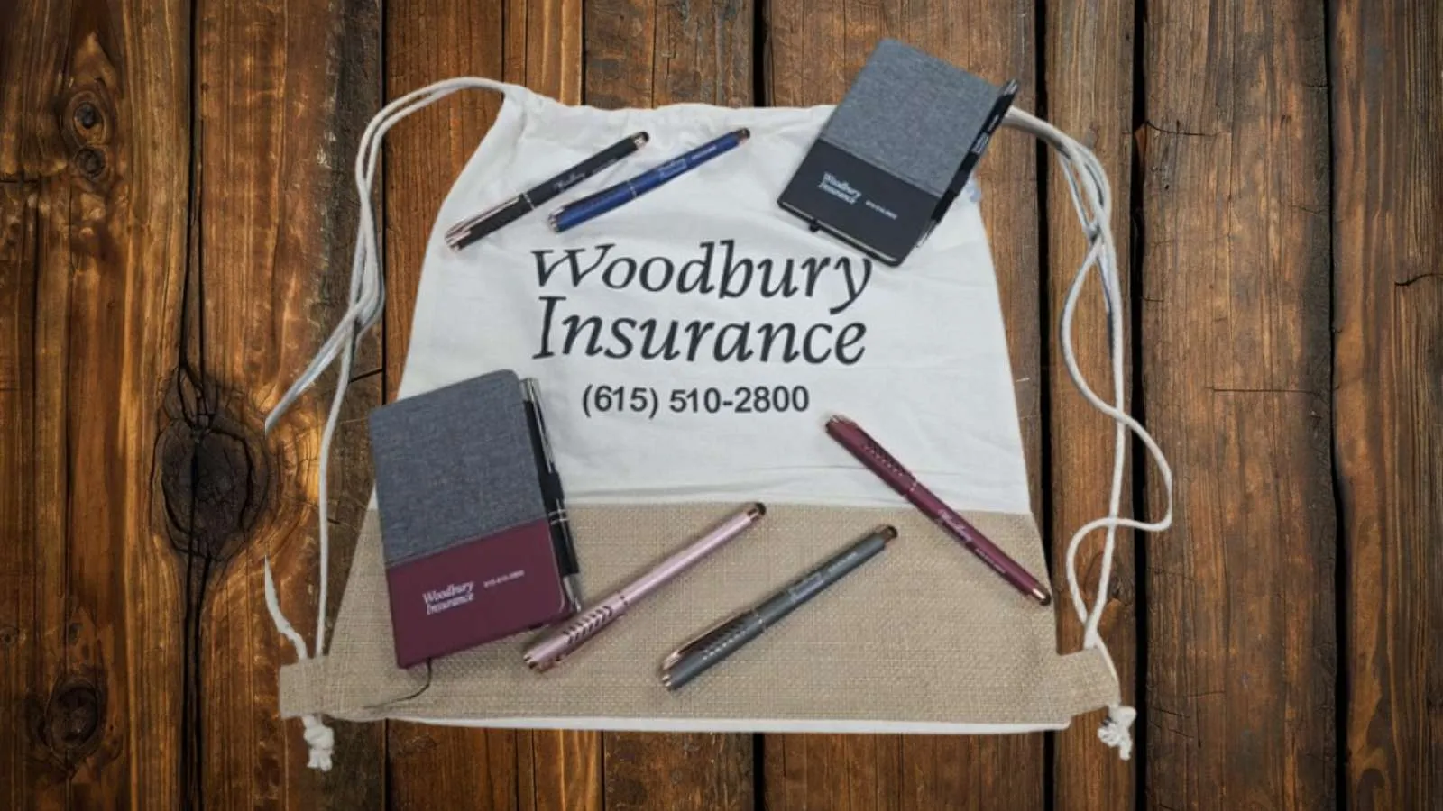 Woodbury Insurance drawstring bag with pens and notebooks on wooden surface showing phone number