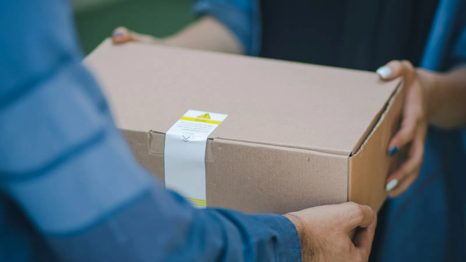 Person handing over a sealed cardboard package to a woman wearing a black scarf outdoors.
