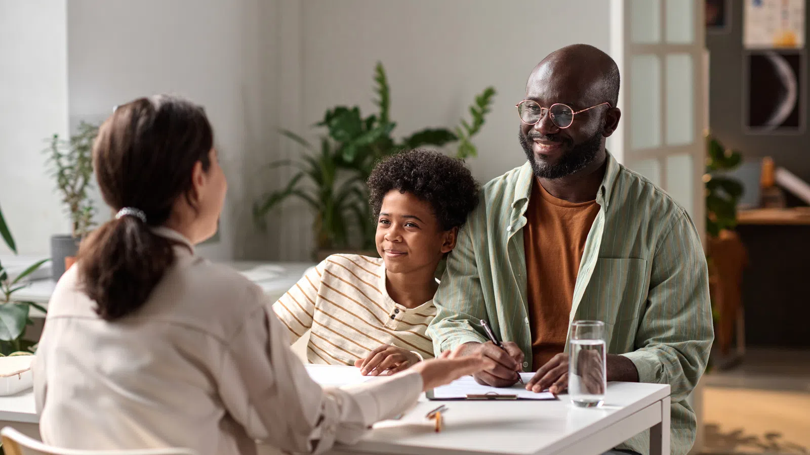 Father and son smiling and talking with a female professional at a bright office desk with plants in the background