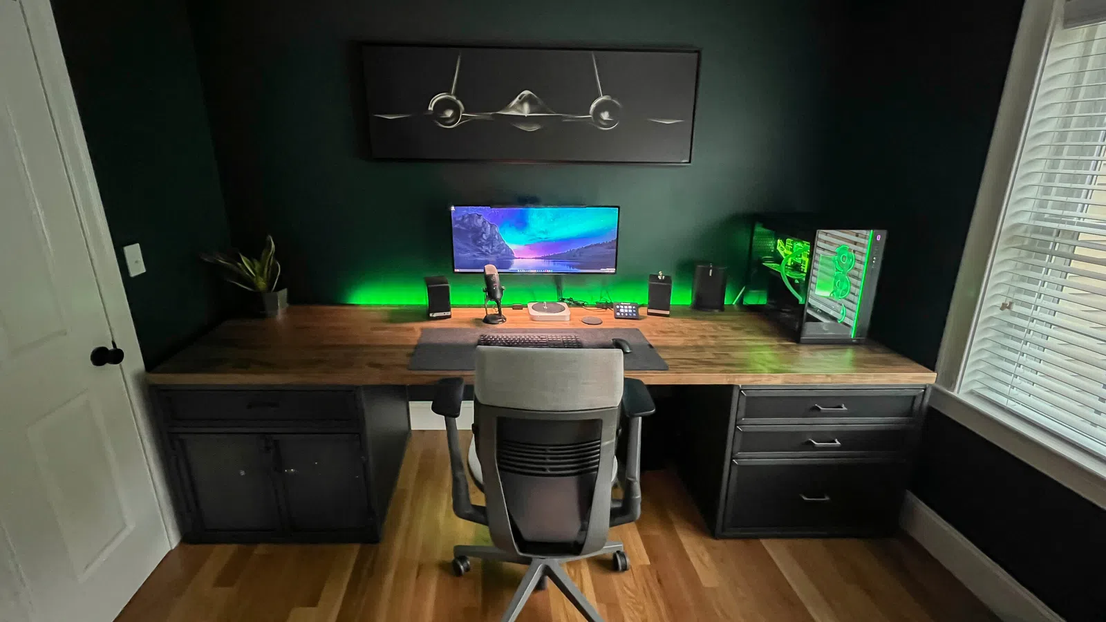 Modern home office setup with a wooden desk, ergonomic chair, computer with green LED lights, and airplane wall art.
