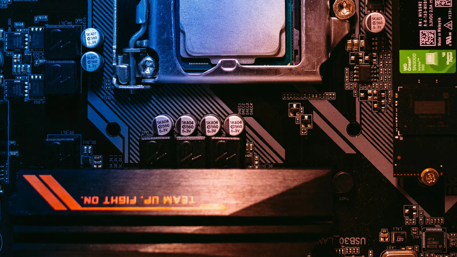 Close-up of a computer motherboard featuring a CPU socket, RAM sticks, and electronic components under blue lighting.