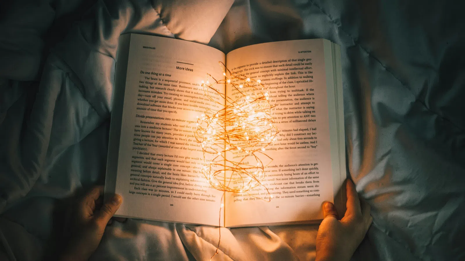 Hands hold an open book with glowing fairy lights resting on soft bed sheets creating a cozy reading scene