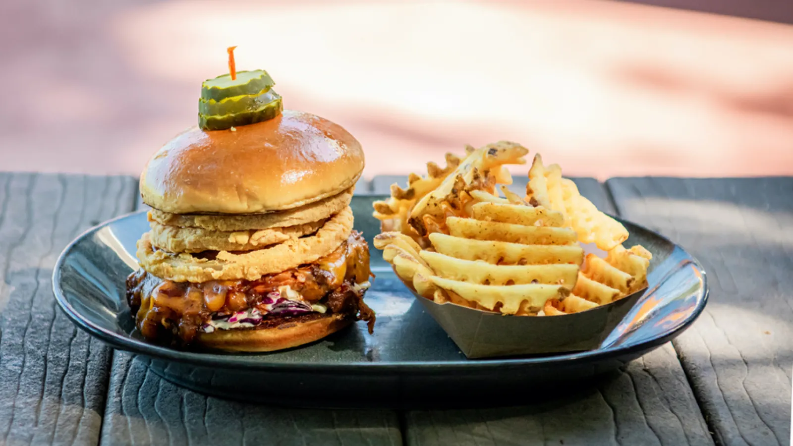 BBQ pulled pork sandwich with fried onion rings and waffle fries served on a black plate outdoors.