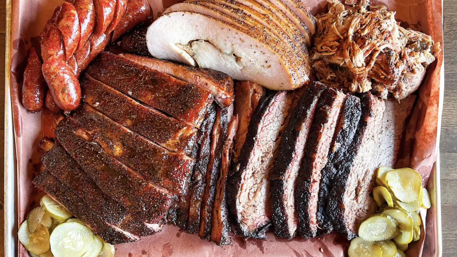 Tray of smoked barbecue meats including ribs, sausage, brisket, turkey, pulled pork, and pickles on parchment paper.