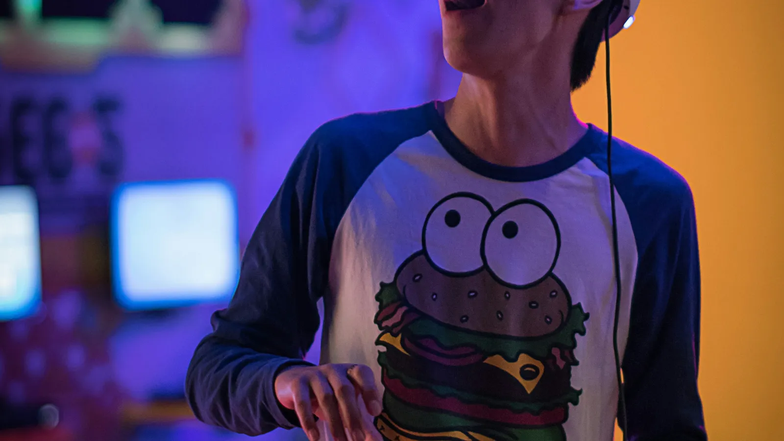 Young man in a cartoon burger shirt using a glowing VR headset in a colorful gaming room at night.