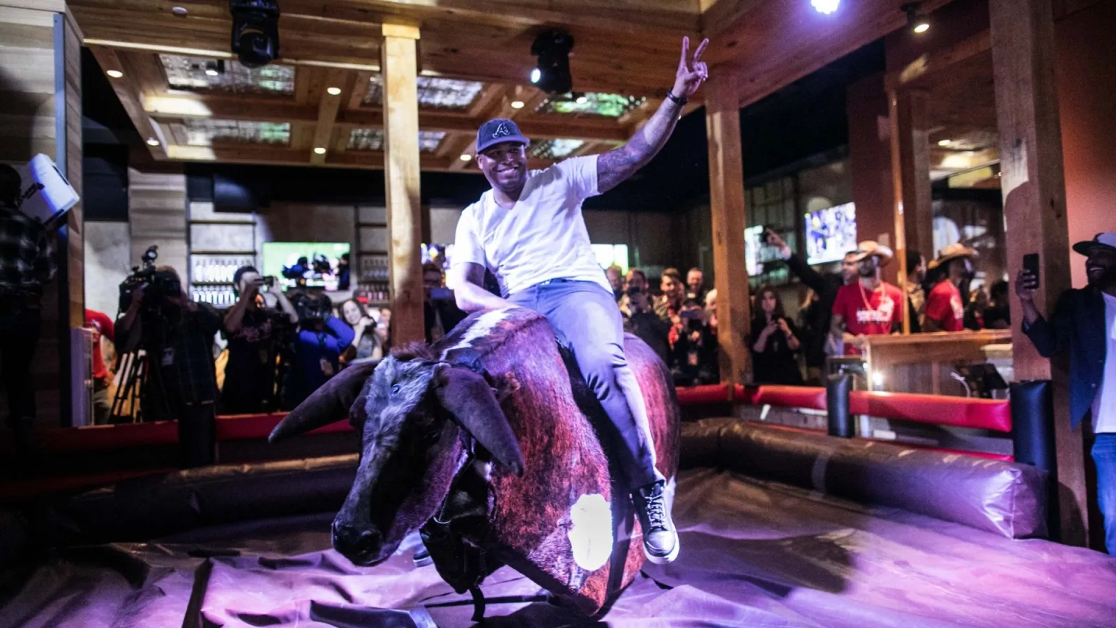 Man in white shirt and baseball cap rides mechanical bull indoors while crowd watches and takes photos.