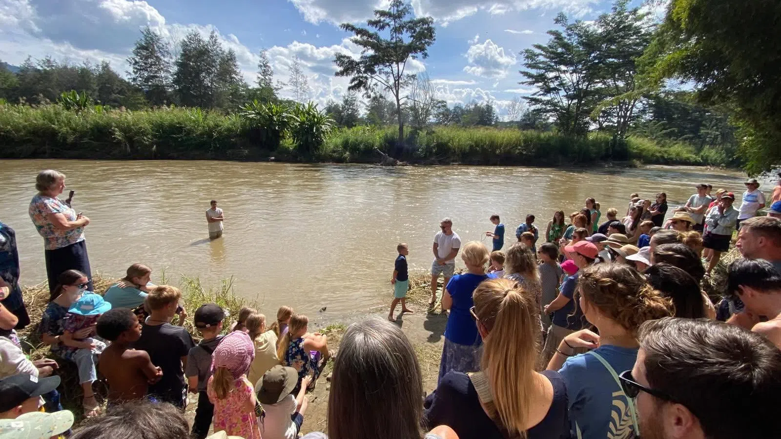 Large group of people gathered by a riverbank watching a man stand in the water under a sunny sky with clouds.