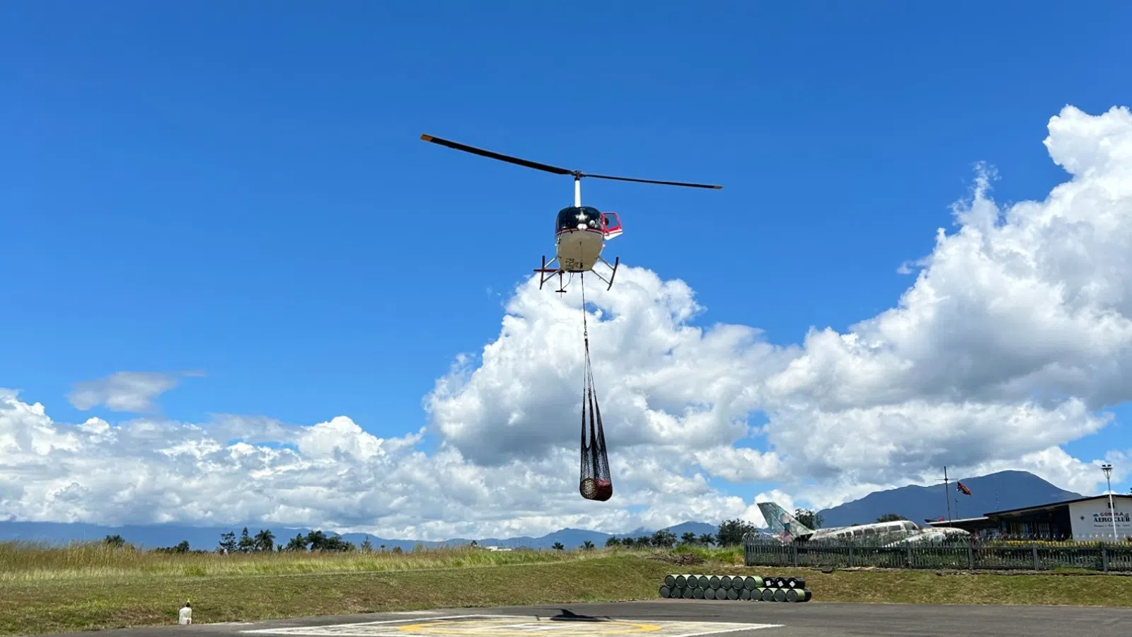 Helicopter carrying a suspended load over a helipad with mountains and blue sky in the background.
