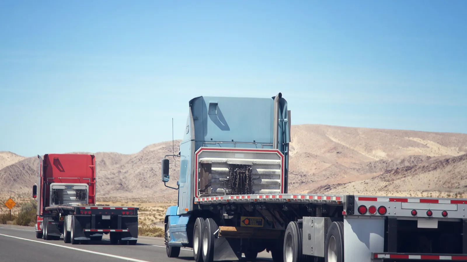 Two semi-trucks driving on a desert highway with mountains in the background under a clear blue sky.