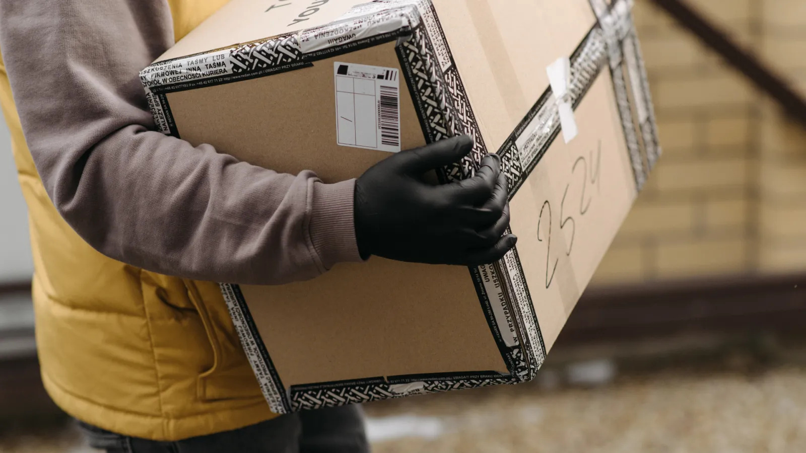 Person wearing black gloves holding a sealed cardboard box with barcode and handwritten numbers outdoors.