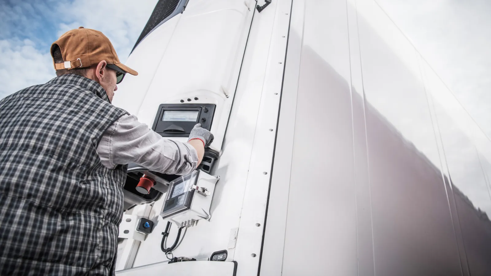 Man in cap and plaid vest operating control panel on a large white refrigerated truck outdoors.