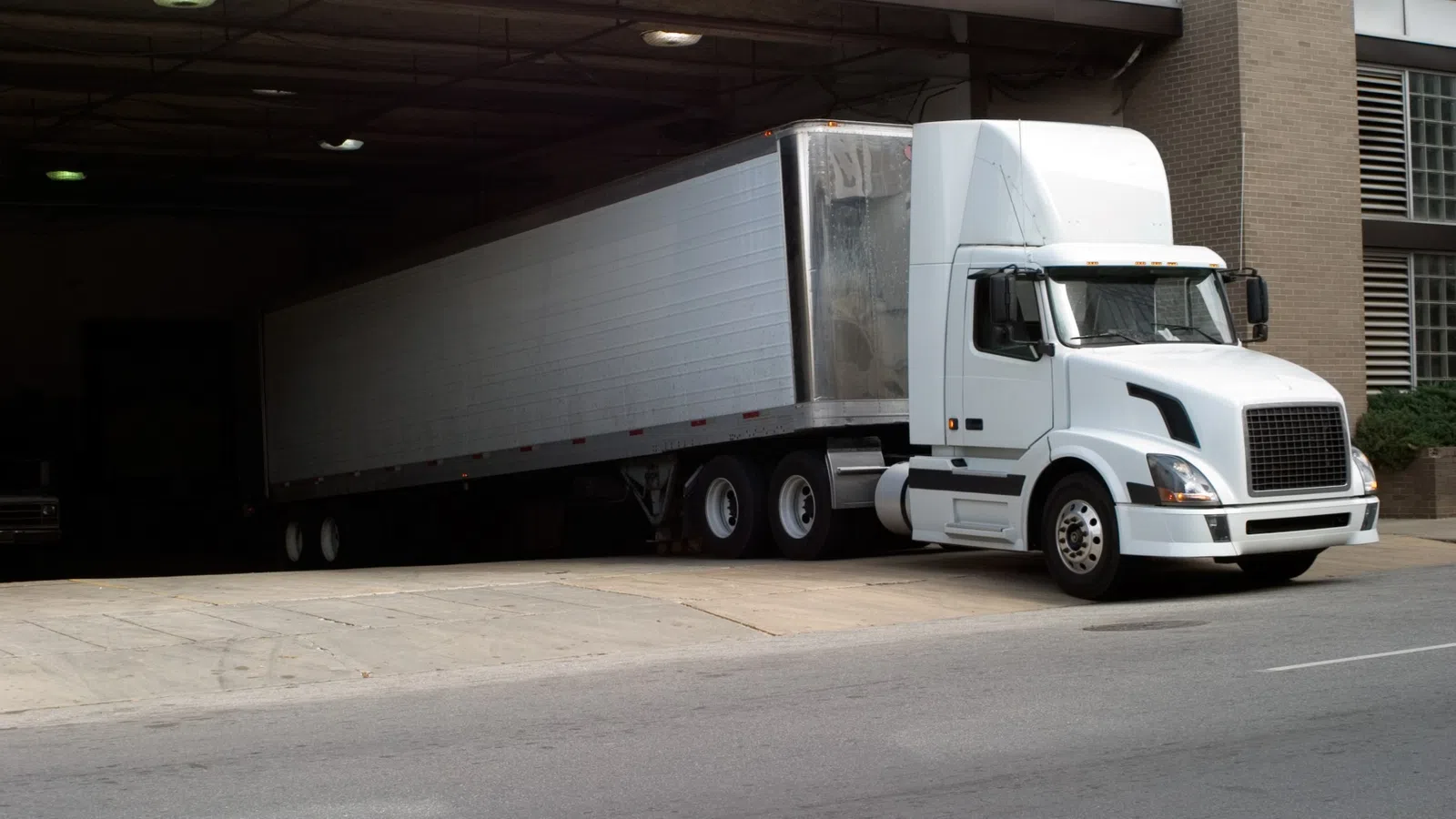White semi-truck with large trailer parked partially under a building overhang near a city street.