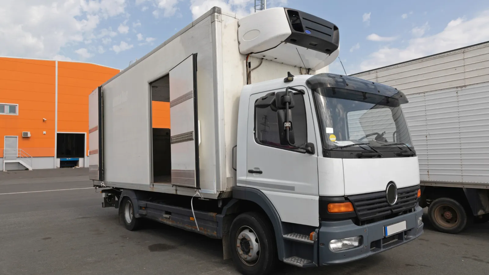 White refrigerated delivery truck with open side door parked near orange warehouse on clear day.