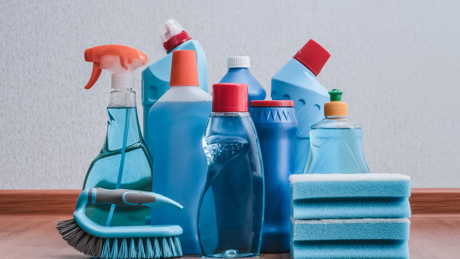 Assortment of blue and red cleaning bottles, brushes, and sponges arranged on wooden floor against white wall.