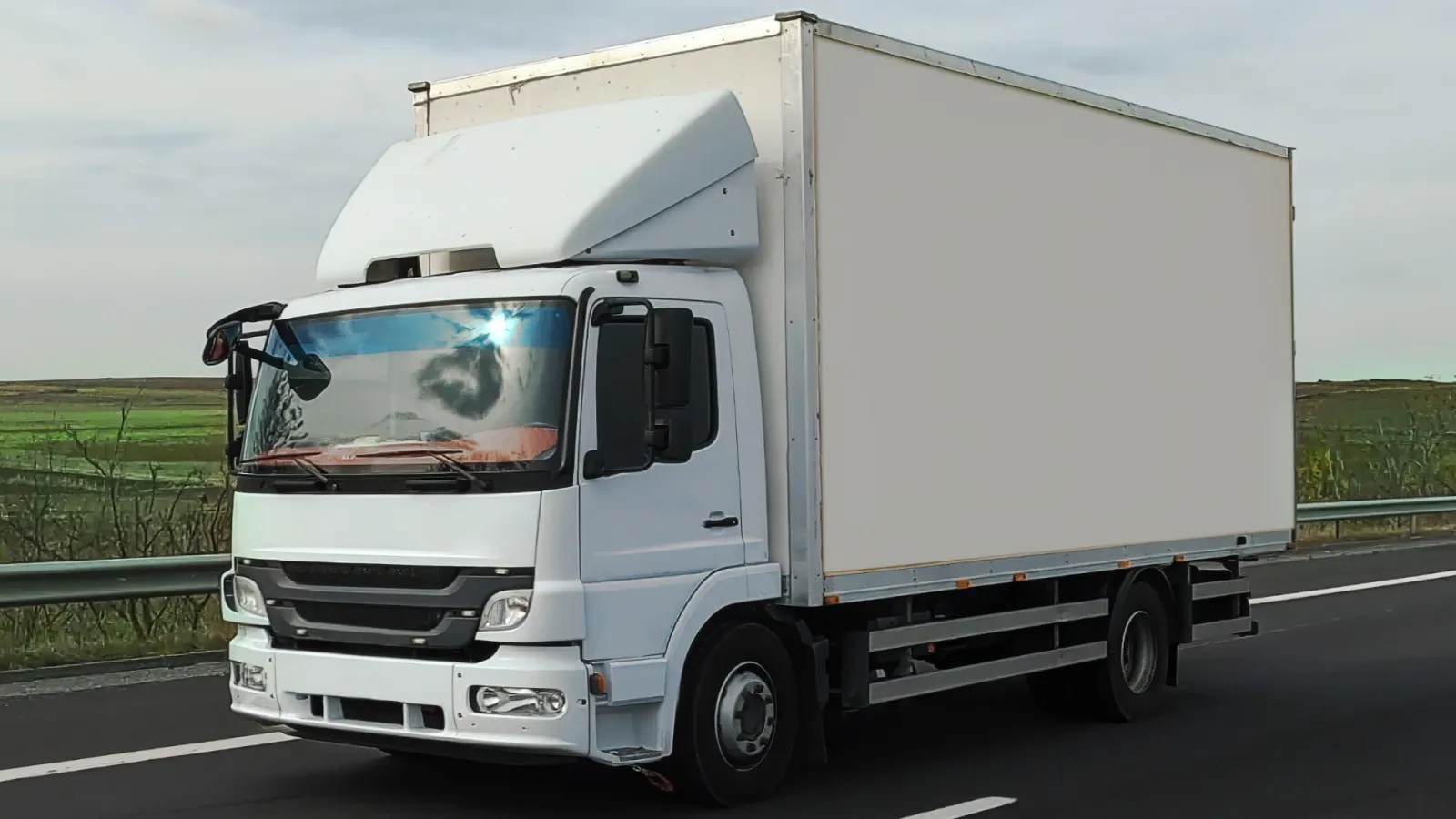 White delivery truck with blank cargo box driving on highway against a cloudy sky and green fields.