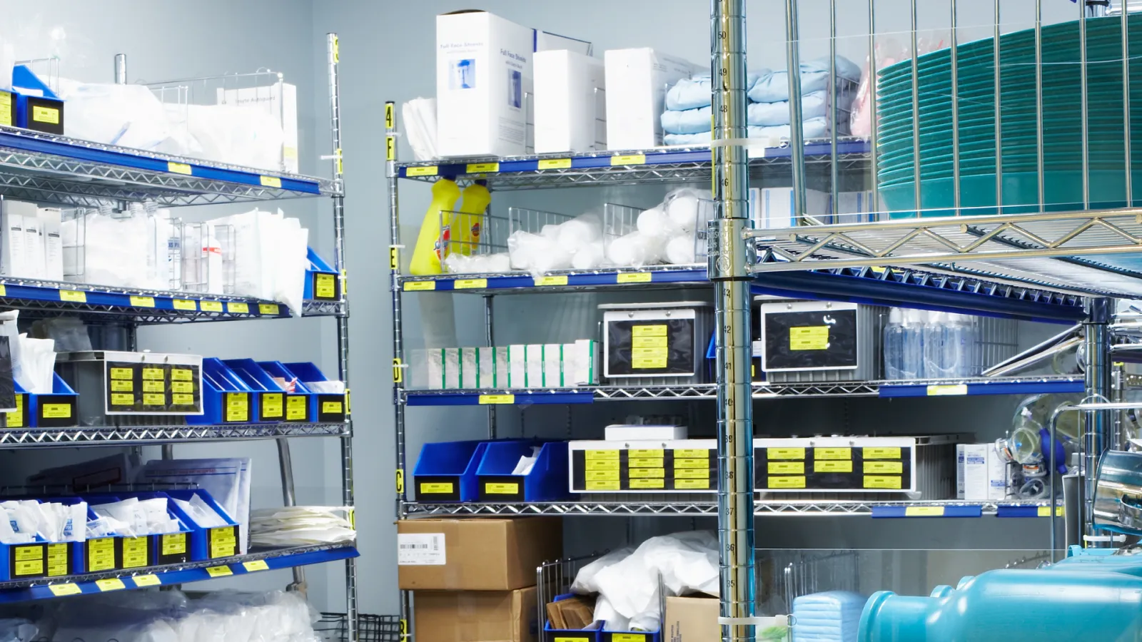 Organized medical supply room with shelves holding boxes, containers, and sterilization equipment in a clinical setting.