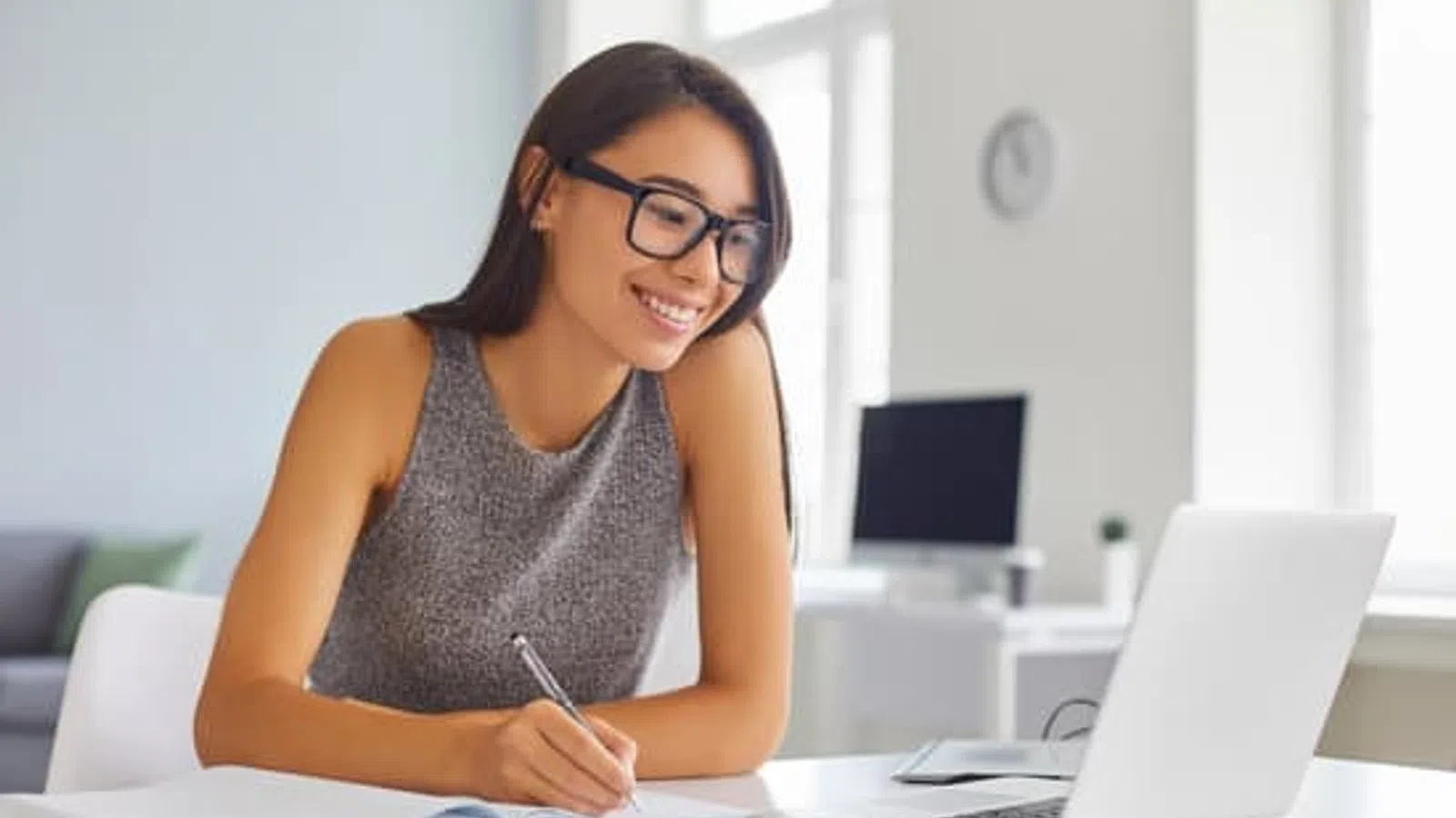 Young woman with glasses smiling while writing in notebook and using laptop in bright modern office.