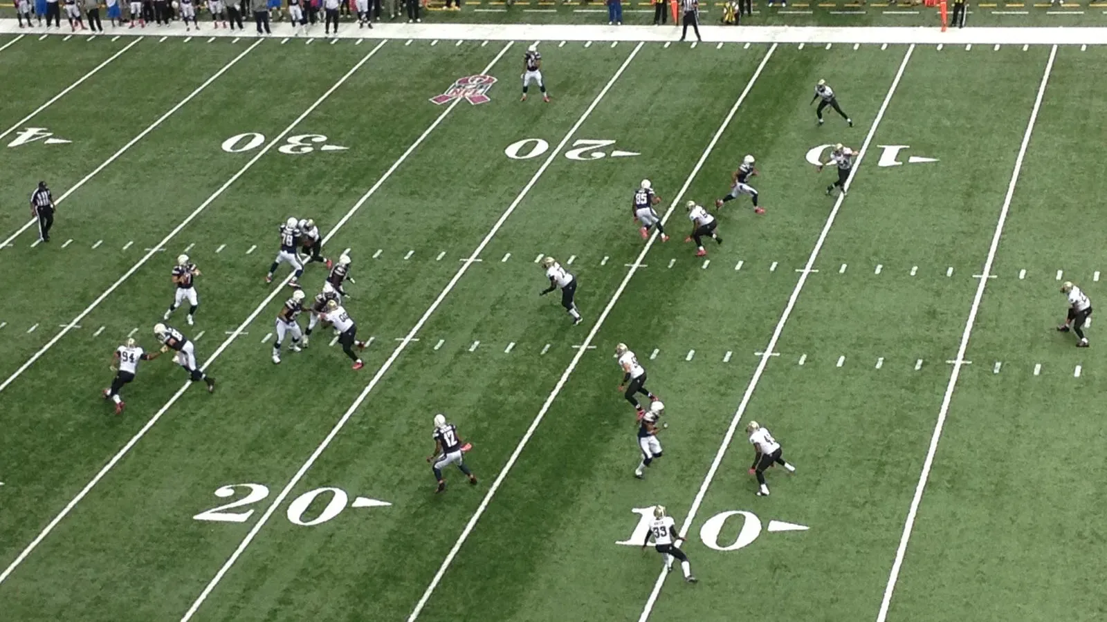 Aerial view of American football game with offense and defense players on green field and spectators in stands.
