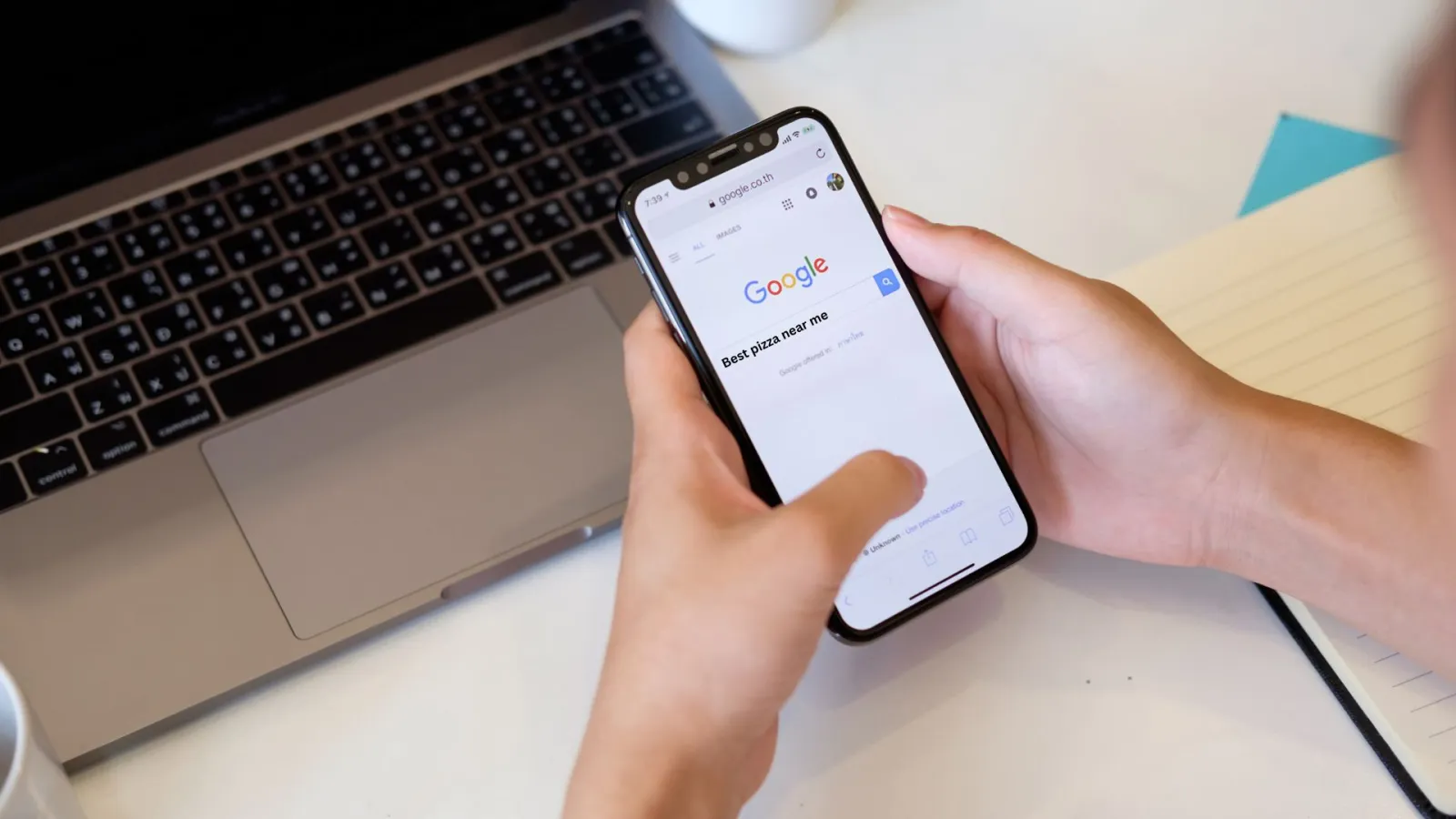 Person holding smartphone searching for best pizza near me on Google with laptop and notebook nearby