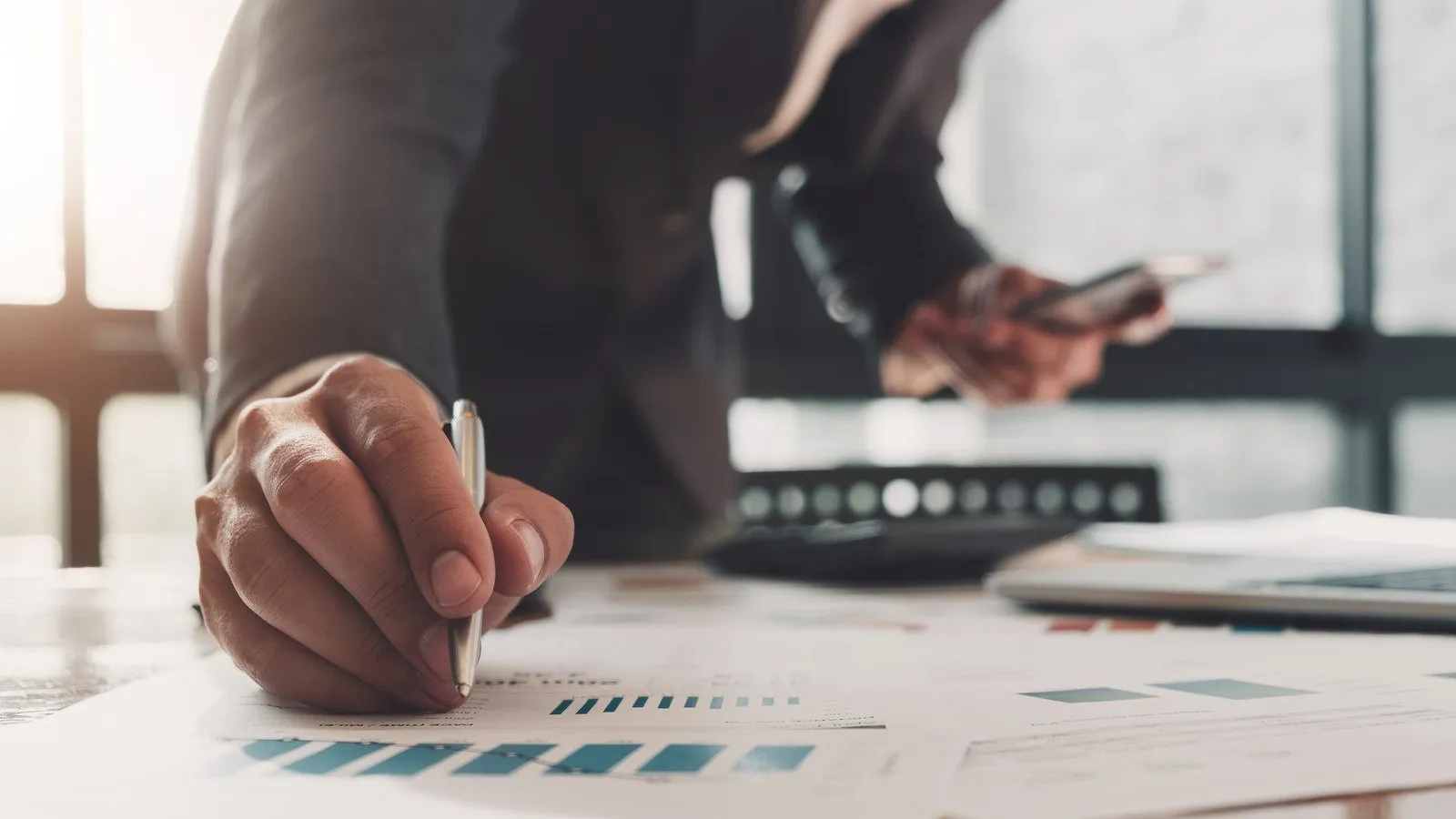 Businessperson analyzing financial charts and data with pen and smartphone in office setting with natural light