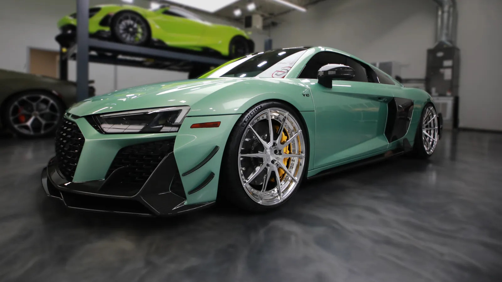 Mint green Audi R8 sports car with custom wheels displayed in a modern showroom with another car on a lift.