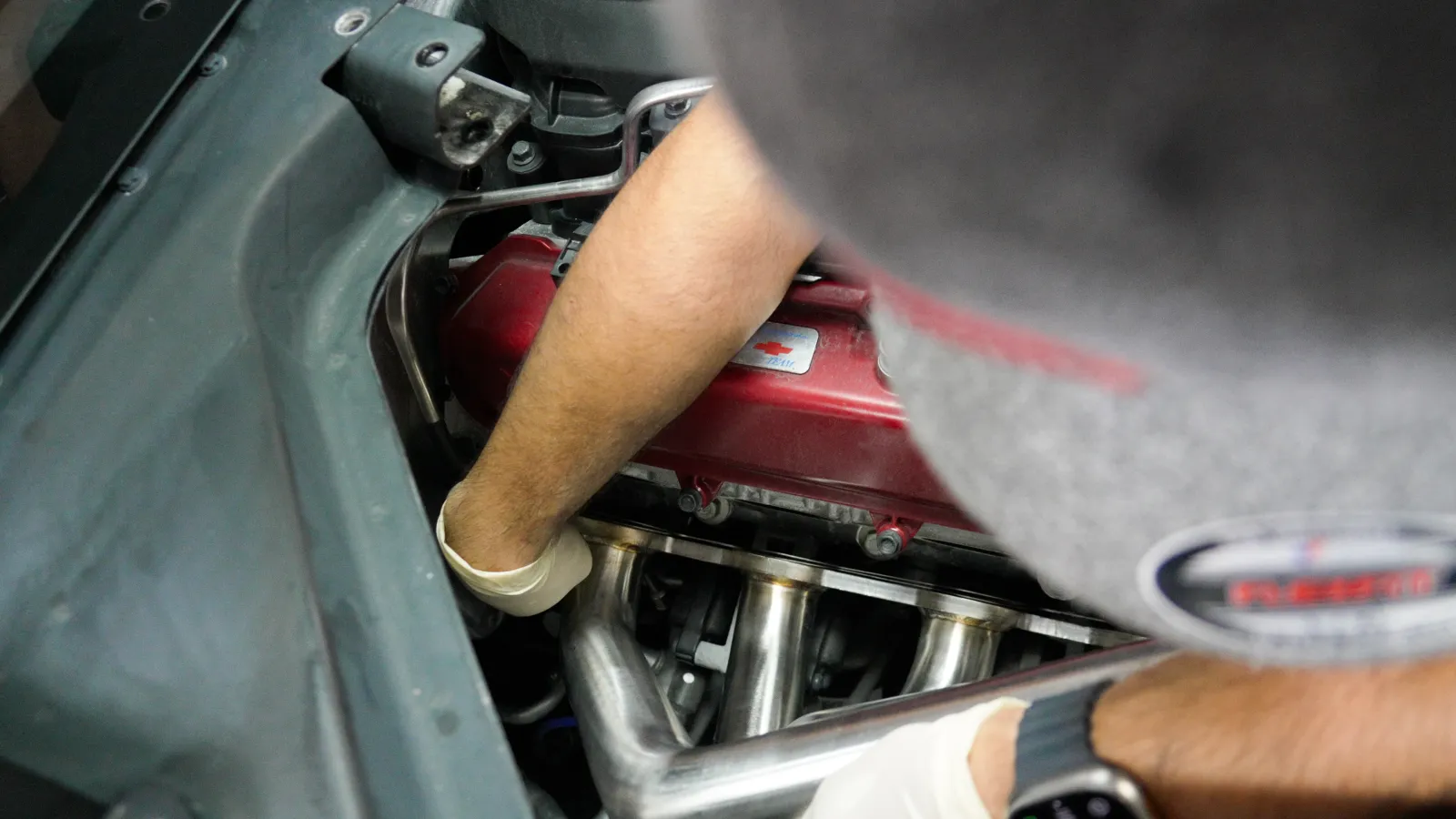 Mechanic wearing gloves working on a red car engine with metal exhaust headers inside an engine bay.