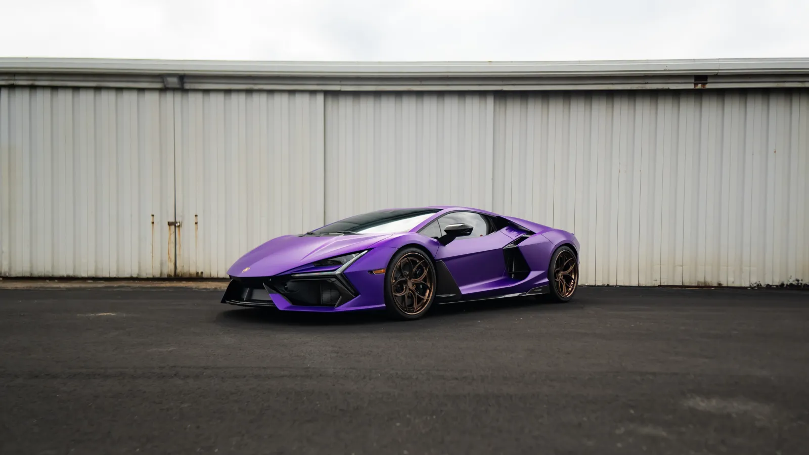 Matte purple sports car with bronze wheels parked on black asphalt against white metal wall.