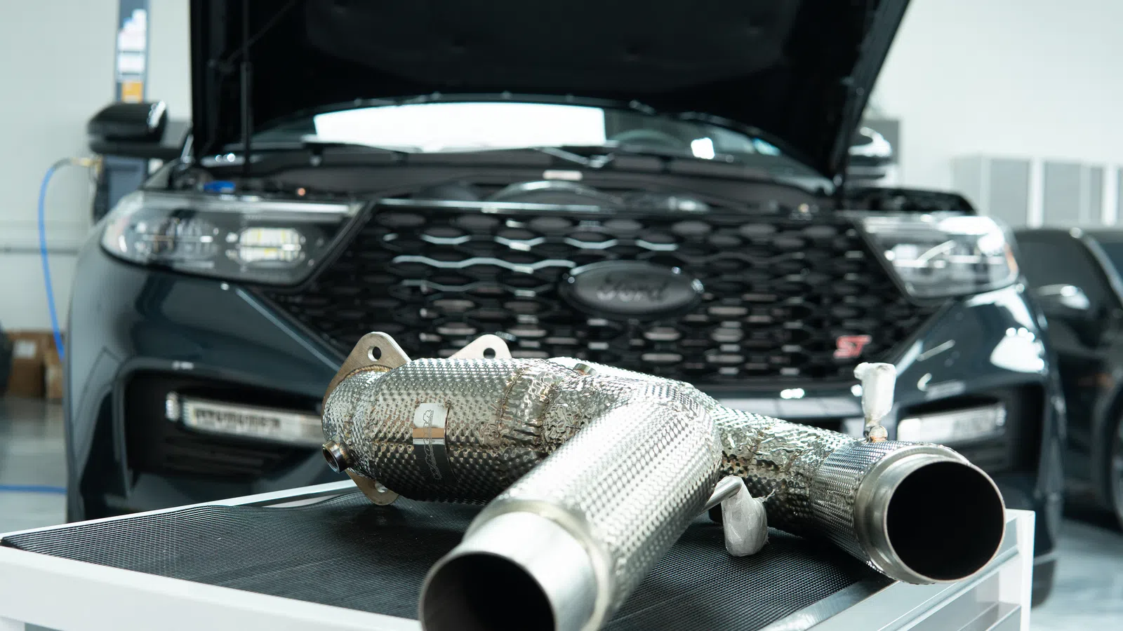 Metal exhaust pipe parts on table with a black car and open hood in the background inside a workshop.