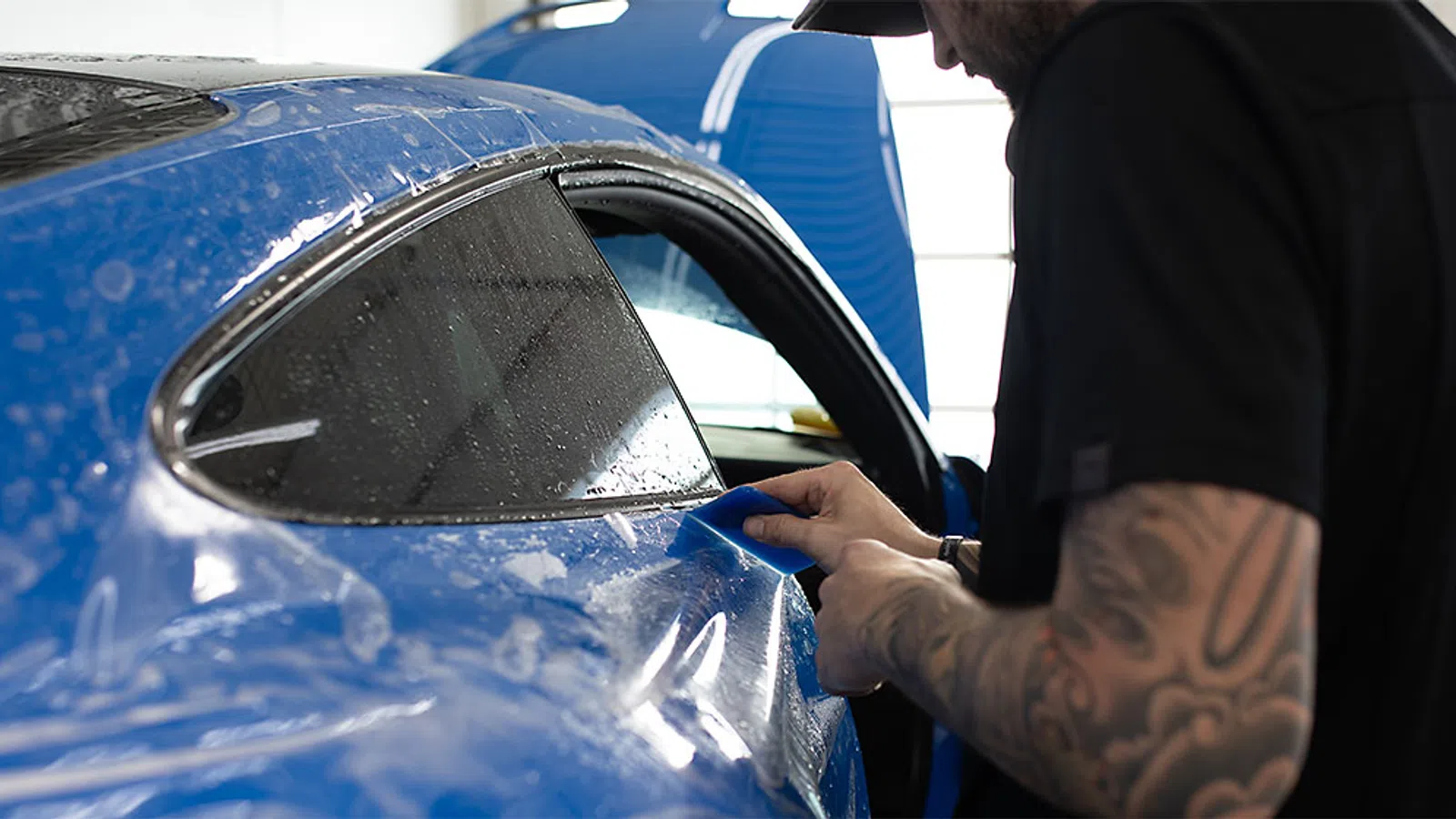 Man applying protective film to blue sports car window in a professional garage setting.