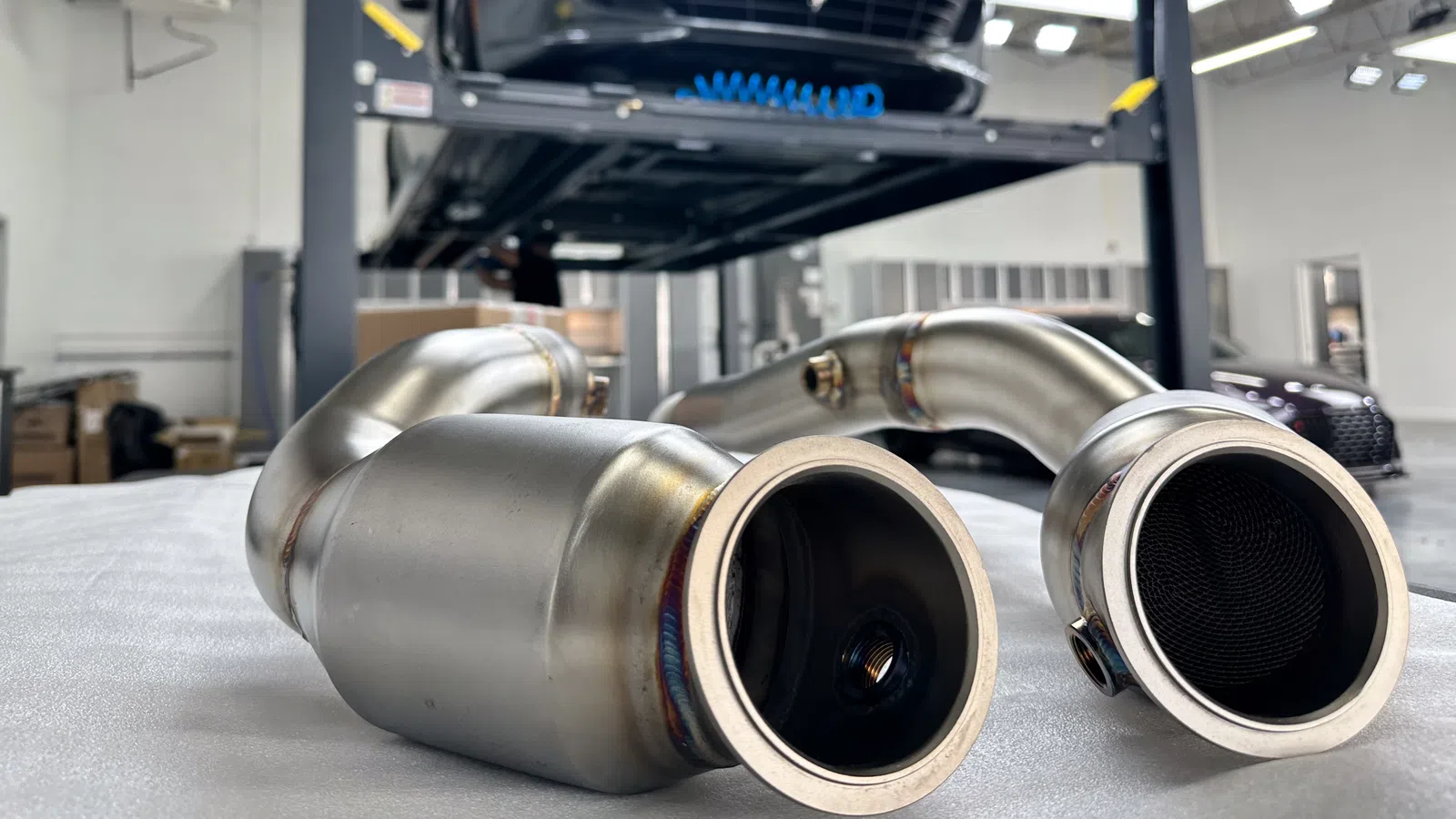 Close-up of stainless steel exhaust pipes with a black sports car on a lift in a garage workshop.