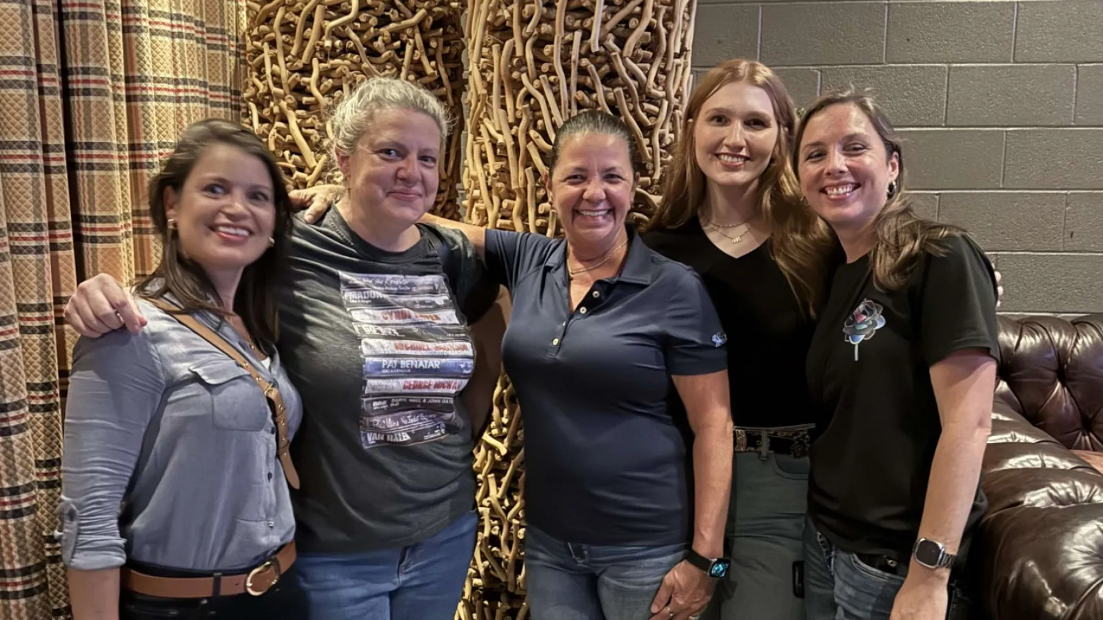 Group of five smiling adults posing indoors near decorative wicker furniture and potted plants under warm lighting