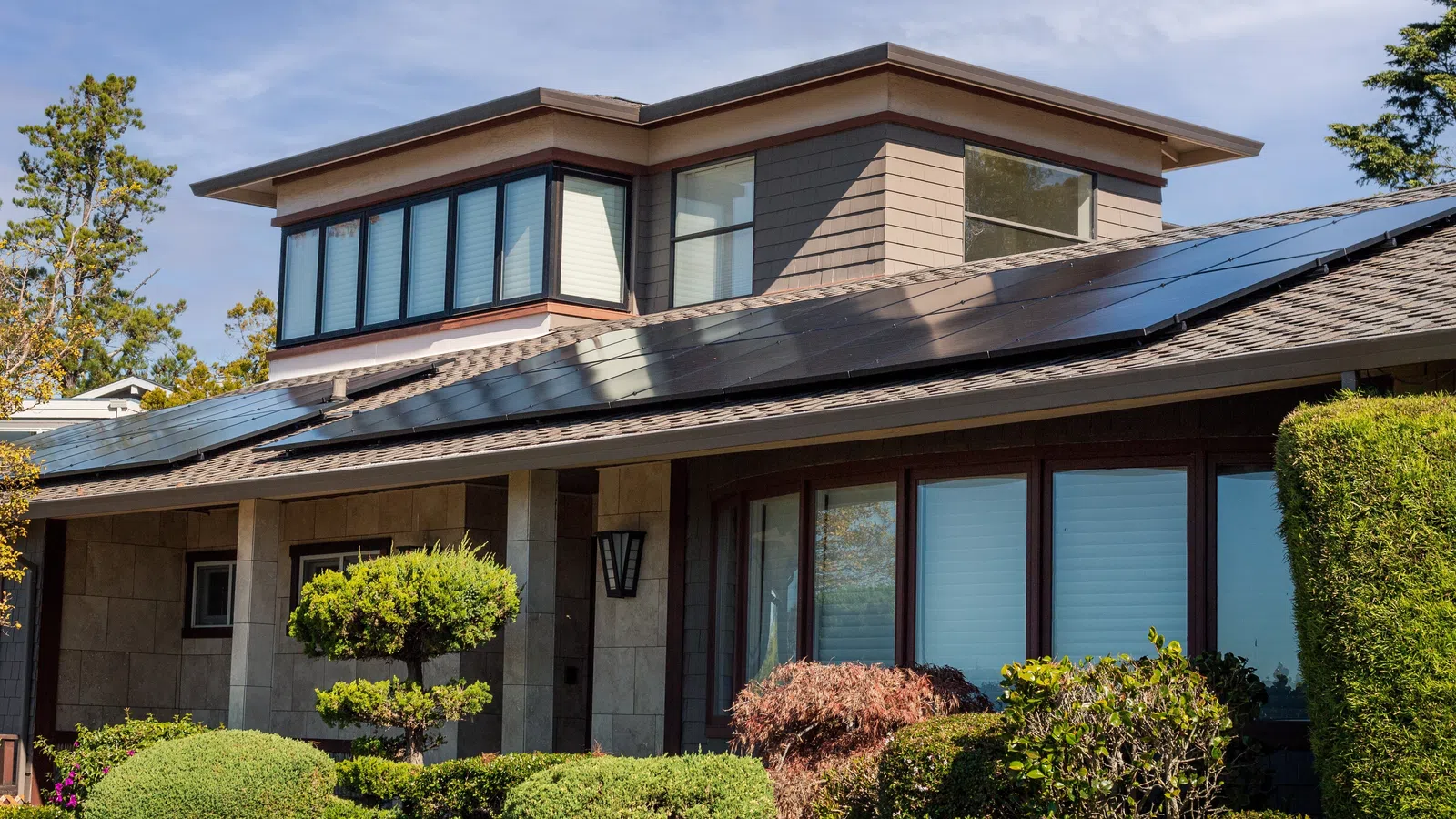 Modern house with solar panels on the roof surrounded by green shrubs and trees under a clear sky.