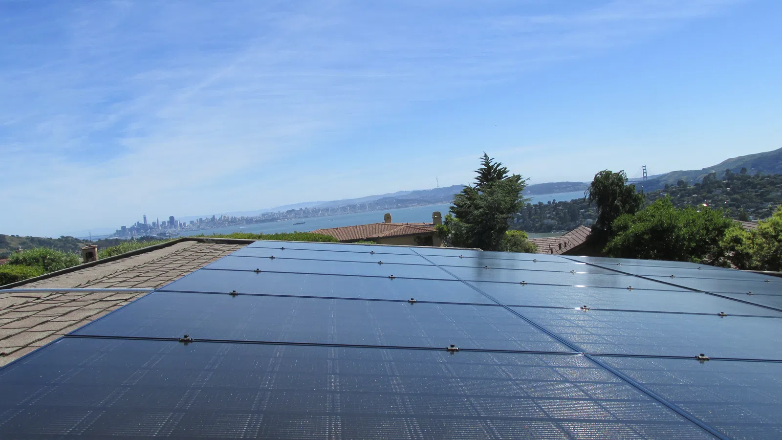 Solar panels installed on a rooftop with San Francisco skyline and Golden Gate Bridge in the background under blue sky.