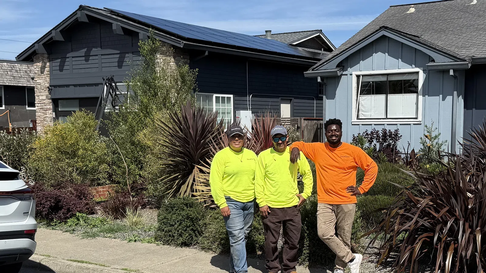 Three workers in bright safety shirts standing on a sidewalk by houses with garden plants and solar panels on the roof.