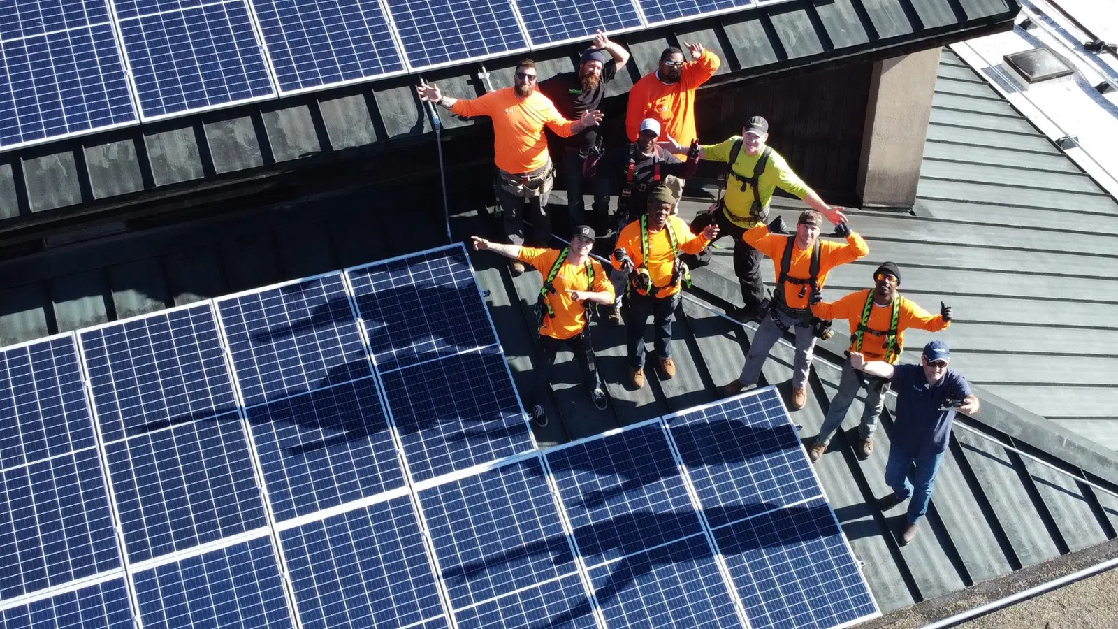 Group of workers in safety gear standing on roof with installed solar panels, waving at the camera.