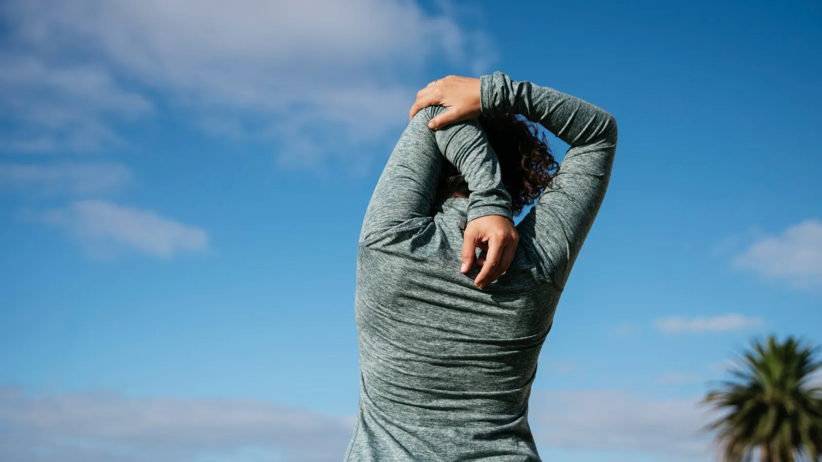 Person in gray hoodie stretching arms overhead outdoors with blue sky and palm tree in background